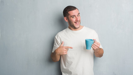 Young caucasian man over grey grunge wall driking a cup of coffee very happy pointing with hand and finger