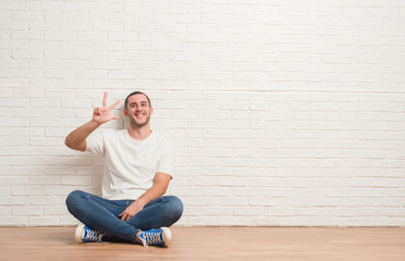 Young caucasian man sitting on the floor over white brick wall showing and pointing up with fingers number three while smiling confident and happy.