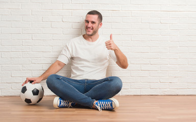 Young caucasian man sitting over white brick wall holding soccer football ball happy with big smile doing ok sign, thumb up with fingers, excellent sign