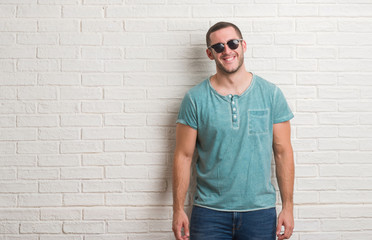 Young caucasian man standing over white brick wall wearing sunglasses with a happy face standing and smiling with a confident smile showing teeth