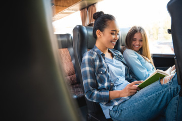 laughing multiethnic female friends reading book during trip on travel bus