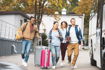 selective focus of smiling multiethnic tourists with wheeled bags and rugby ball running near travel bus at city street