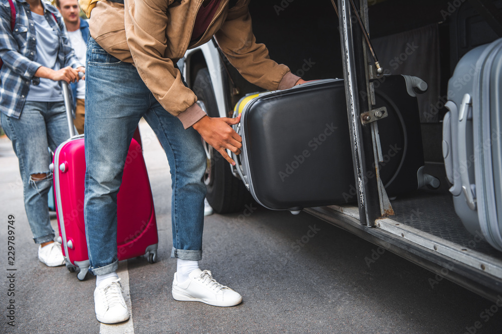 Wall mural cropped image of man putting wheeled bag into travel bus while his friends standing behind at street