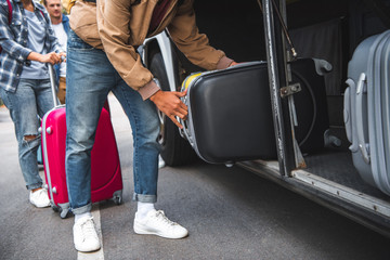 cropped image of man putting wheeled bag into travel bus while his friends standing behind at street