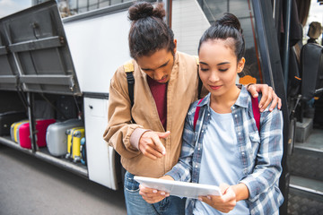 multiracial young man pointing by finger at digital tablet to asian girlfriend near travel bus at street