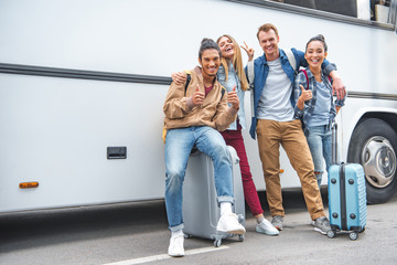 smiling multiethnic friends with wheeled bags doing thumbs up and peace gestures near travel bus