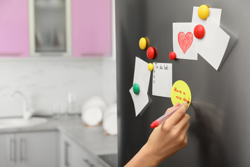Woman writing message on note stuck to refrigerator door at home, closeup