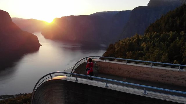 Aerial view. Tourist woman with norwegian flag and photo camera enjoying fjord Aurlandsfjord view from Stegastein viewing point. National tourist route Aurlandsfjellet, Norway