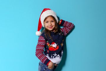 Cute little girl in Christmas sweater and hat on color background