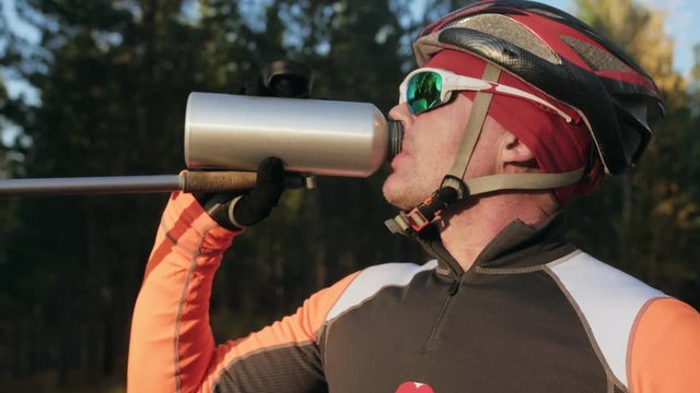 Training an athlete on the roller skaters. Biathlon ride on the roller skis with ski poles, in the helmet. Autumn workout. Roller sport. Adult man riding on skates. The athlete drinks water from a