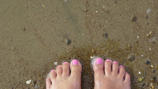 Top view of beautiful female barefeet legs with professional pink painted toenails. Pink color pedicure. White tanned woman standing and relaxing on sandy beach at seashore on summer day.