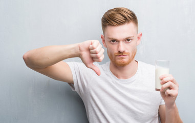 Young redhead man over grey grunge wall drinking a glass of milk with angry face, negative sign showing dislike with thumbs down, rejection concept