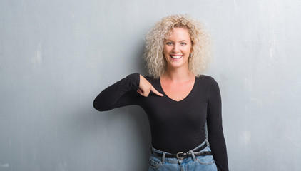 Young blonde woman with curly hair over grunge grey background with surprise face pointing finger to himself