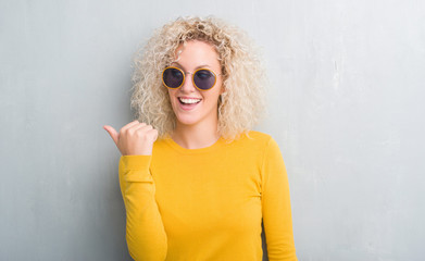 Young blonde woman with curly hair over grunge grey background smiling with happy face looking and pointing to the side with thumb up.