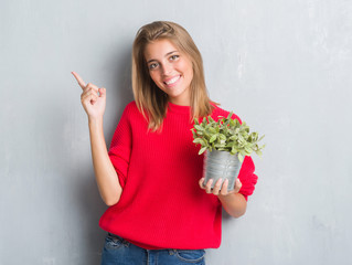 Beautiful young woman over grunge grey wall holding plant pot very happy pointing with hand and finger to the side