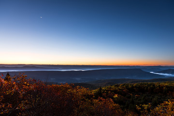 Bear rocks overlook during sunrise, dawn, moon in autumn with rocky landscape in Dolly Sods, West Virginia with orange foliage trees, blue, yellow sky, layered mountains, hills