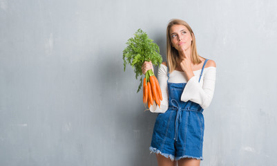 Beautiful young woman over grunge grey wall holding fresh carrots serious face thinking about...
