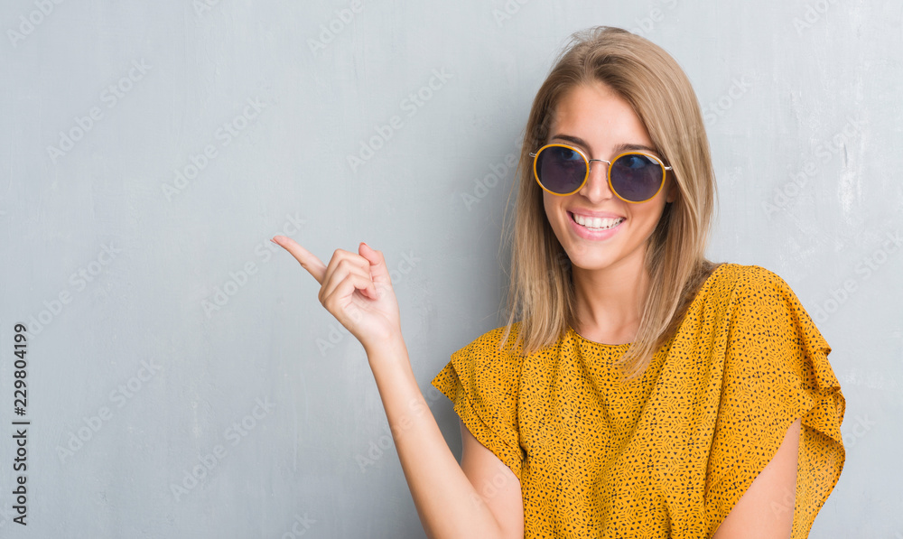 Wall mural Beautiful young woman standing over grunge grey wall wearing retro sunglasses very happy pointing with hand and finger to the side