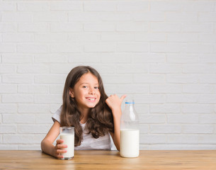 Young hispanic kid sitting on the table drinking a glass of milk pointing and showing with thumb up to the side with happy face smiling