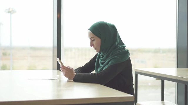 Pretty muslim woman in hijab sitting at the table and uses the phone,slow mo