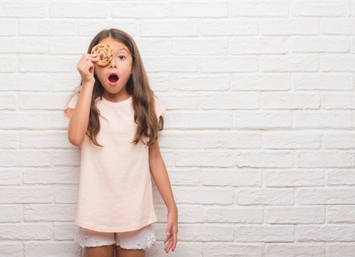 Young hispanic kid over white brick wall eating chocolate chips cooky scared in shock with a surprise face, afraid and excited with fear expression