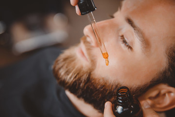Means for caring beard oil, barber puts on face of man.