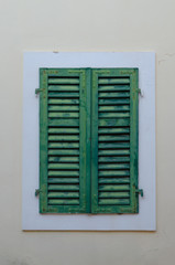 One window with closed green shutters on the background of beige wall.