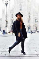 Fashion model walking by the Duomo of Milan looking back wearing a hat and a dark coat and a...