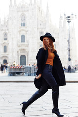 Fashion model walking by the Duomo of Milan wearing a hat and a dark coat in a October morning...