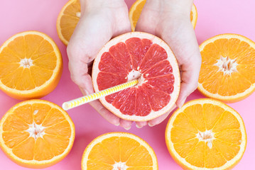 Female hands holding half of a juicy ripe red grapefruit with a straw above oranges cut in half, top view.