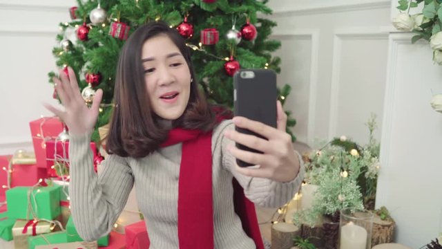 Cheerful happy young Asian woman using smartphone selfie with christmas tree decorate her living room at home in Christmas Festival. Lifestyle woman celebrate Christmas and New year concept.