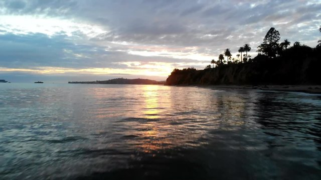 Aerial drone shot at sunset flying low over the water and reflections of the Pacific Ocean surface with the silhouette of California coast cliffs and palm trees in Santa Barbara, California.