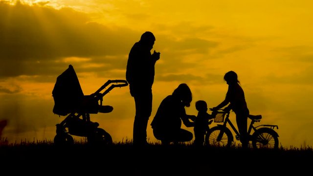 Silhouettes of happy parents having good time with their little children on the outdoor at sunset. Silhouettes of parents walk with small children with a stroller and bicycle.