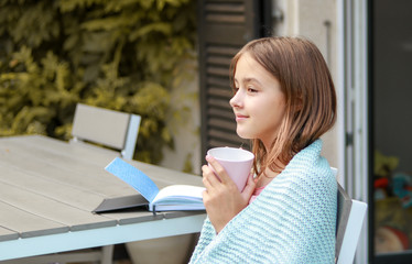 Fototapeta premium Beautiful smiling young girl daydreaming with cup of tea in her hands and book on table outside in autumn garden wrapped into warm knitted blanket. Lagom style. Cozy autumn