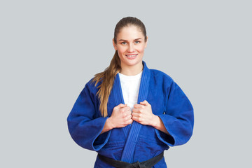 Portrait of happy beautiful athletic karate woman in blue kimono with black belt standing and looking at camera with toothy smile. Japanese martial arts concept. Indoor, studio shot, gray background