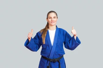Fototapete Kampfkunst Positive athletic karate woman in blue kimono with black belt standing, showing thumbs up and looking at camera with toothy smile. Japanese martial arts concept. Indoor, studio shot, gray background