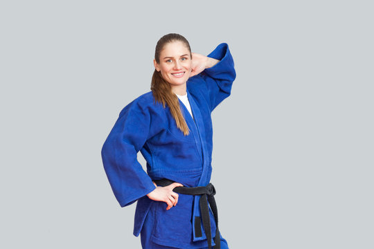 Beautiful happy athletic karate woman in blue kimono with black belt posing and holding hand behind her head are looking at camera. Japanese martial arts concept. Indoor, studio shot, gray background