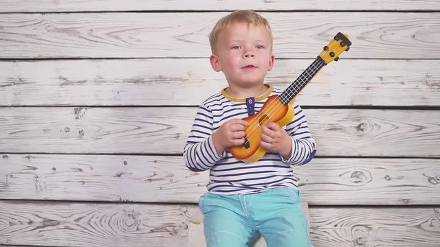One year old boy plays his guitar or ukulele and sings songs, sitting in the wooden room.