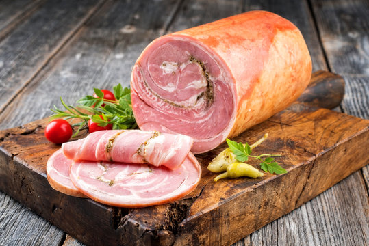 Traditionelle Allgäuer Kalbsrolle Wurst am Stück und aufgeschnitten mit Tomaten und peperoni als closeup auf einem alten rustikalen Schneidebrett 