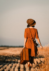 photo of the beautiful young woman with camera and brown suitcase walking in the field