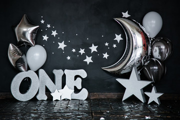 Smashed first birthday white cake with stars and one candle for little baby boy and decorations. Black background. Big silver letters ONE, silver stars and different balloons