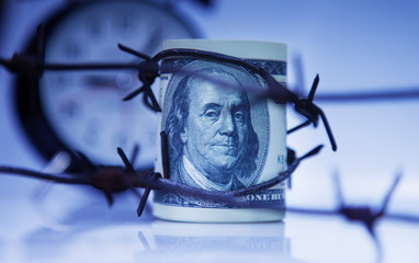 Alarm clock and US Dollar money wrapped in barbed wire as symbol of economic warfare, sanctions and embargo busting