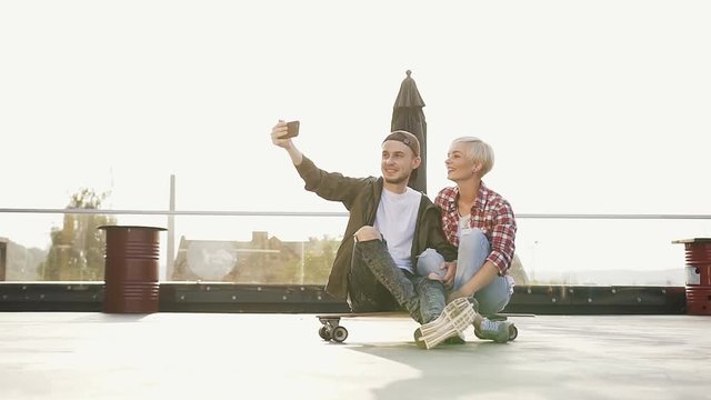 Portrait of young smiling couple dressed in trendy clothes having fun while makes a selfie photo together on the roof of the outdoors