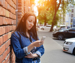 A hipster girl, dressed in urban style, leans against a brick wall with a phone in her hands. The teenager dials a text message in the messenger. Summer sunlight. The sunrise. Communications.