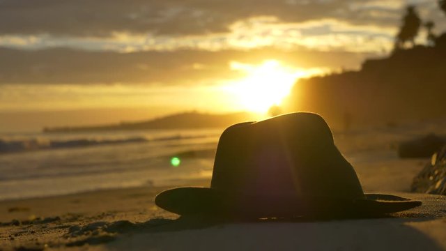 Golden sunset on Butterfly Beach in Montecito, California with a hat in the sand, peaceful ocean waves washing against the shore and people in silhouette walking in the distance.