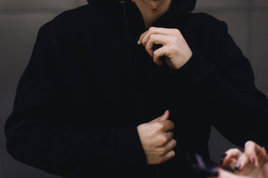 Young Man Zipping Up His Hoodie. Man Dressing Black Sport Clothes. Grey Urban Background. In The Foreground Are Women's Hands With A Telephone.