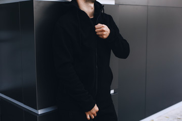 Young man zipping up his hoodie. Man dressing black sport clothes. Grey urban background. 