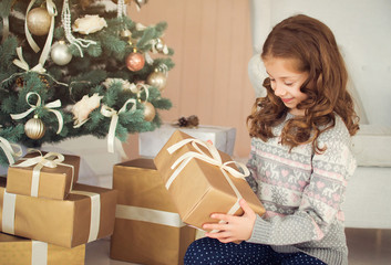 Happy Holidays. Cute little baby opens a Christmas present. Girl happy gifts sitting at the Christmas tree.