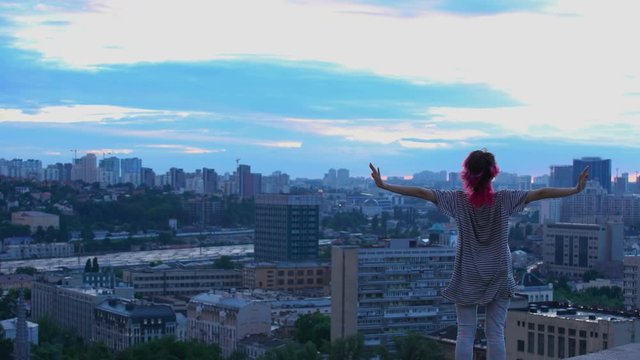 Young female looks at city scape in twilight, pink hair teenager on roof top