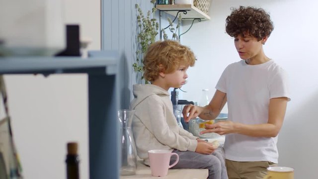 Medium shot of happy mother with short curly hair making peanut butter sandwich and chatting with adorable little boy sitting on kitchen counter and snaking on bread